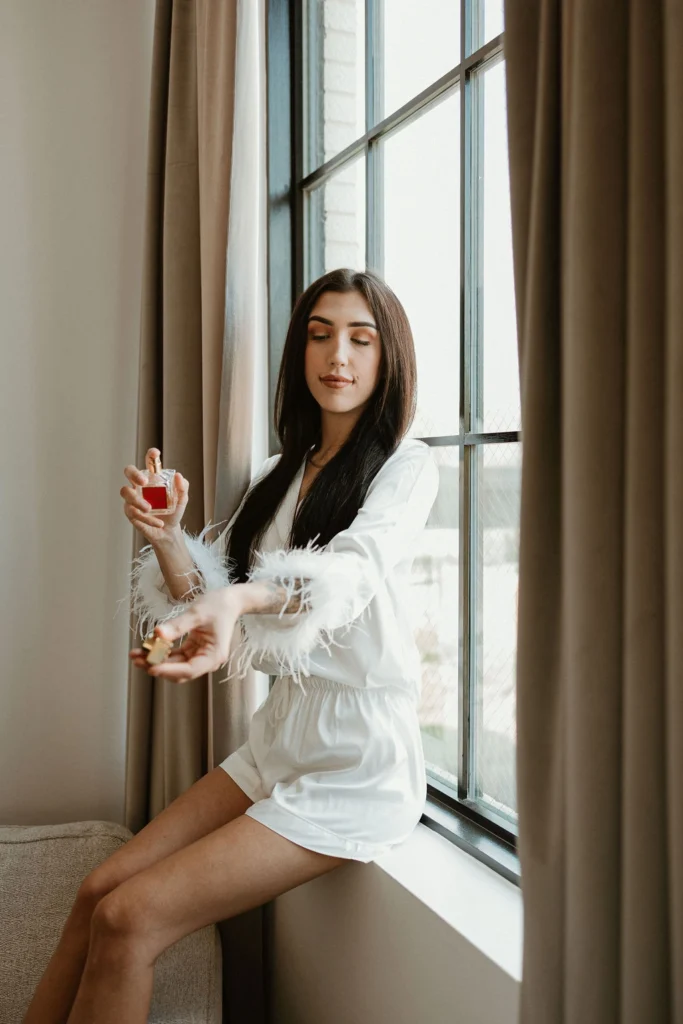 A bride, seated in a window sill, sprays perfume on her wrist