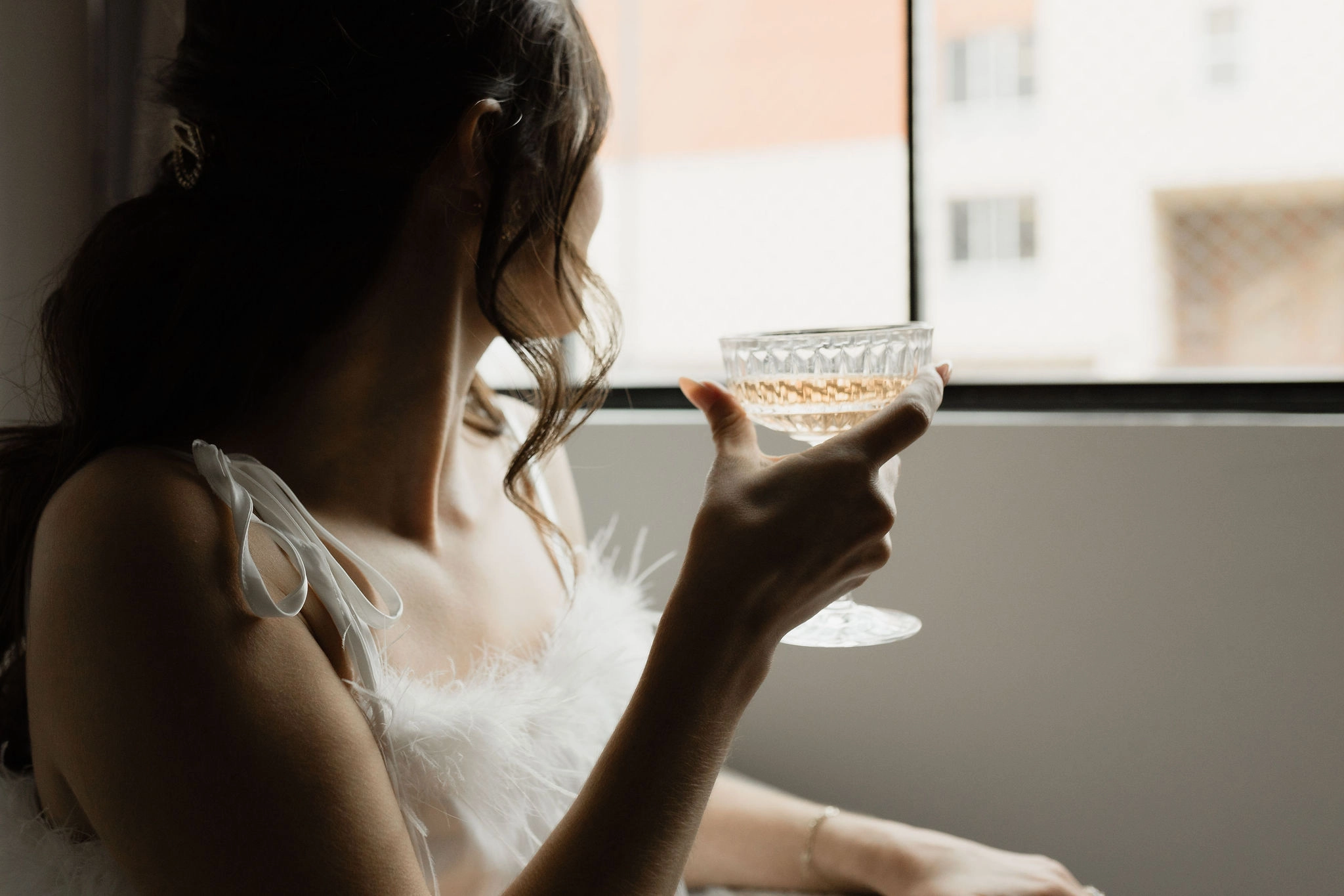 A bride holds a drink in a glass chalice while looking out the window