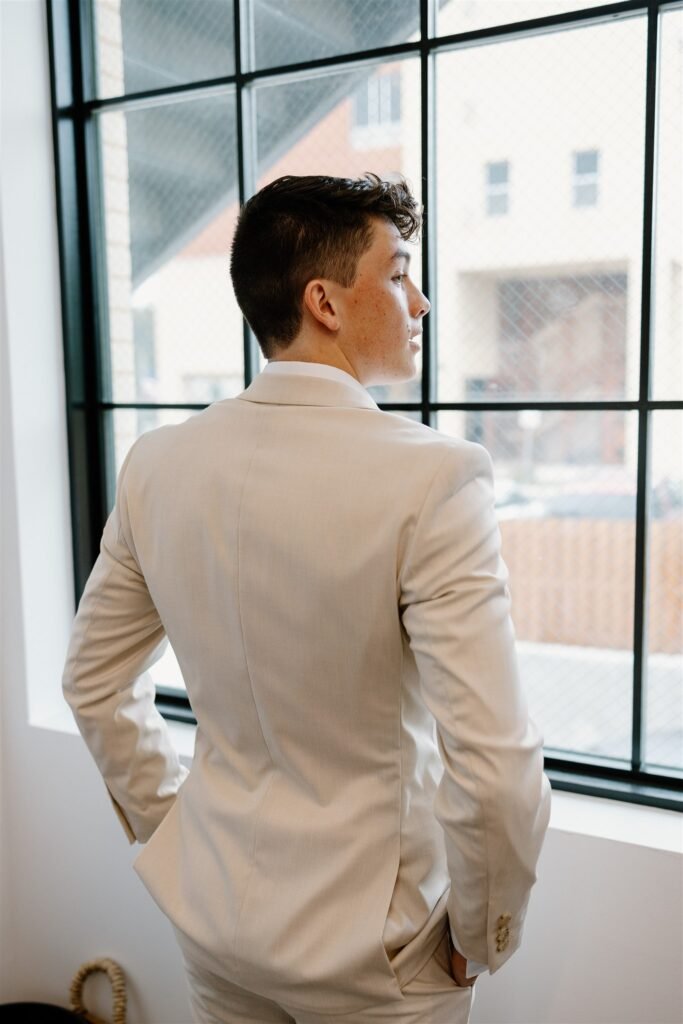 A groom looks out a window. He's dressed in a light colored tuxedo