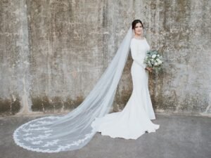 A bride pictured in front of a stone wall. Her veil trails behind her.