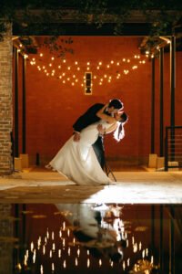 A newly married couple kisses under string lights. Their reflection is seen in a water feature in the foreground