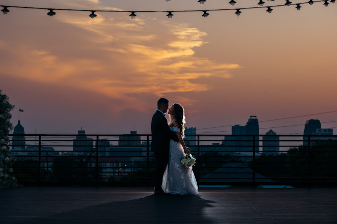 A couple embraces. He's in a tux, she's in a wedding dress. They are overlooking the San Antonio skyline with a beautiful sunset backdrop